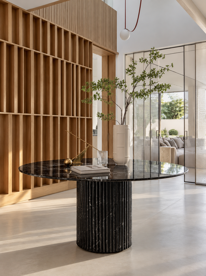 Dominio round dining table in Black Marquina marble on a fluted cylindrical marble base in a bright entrance hall with oak slatted shelving